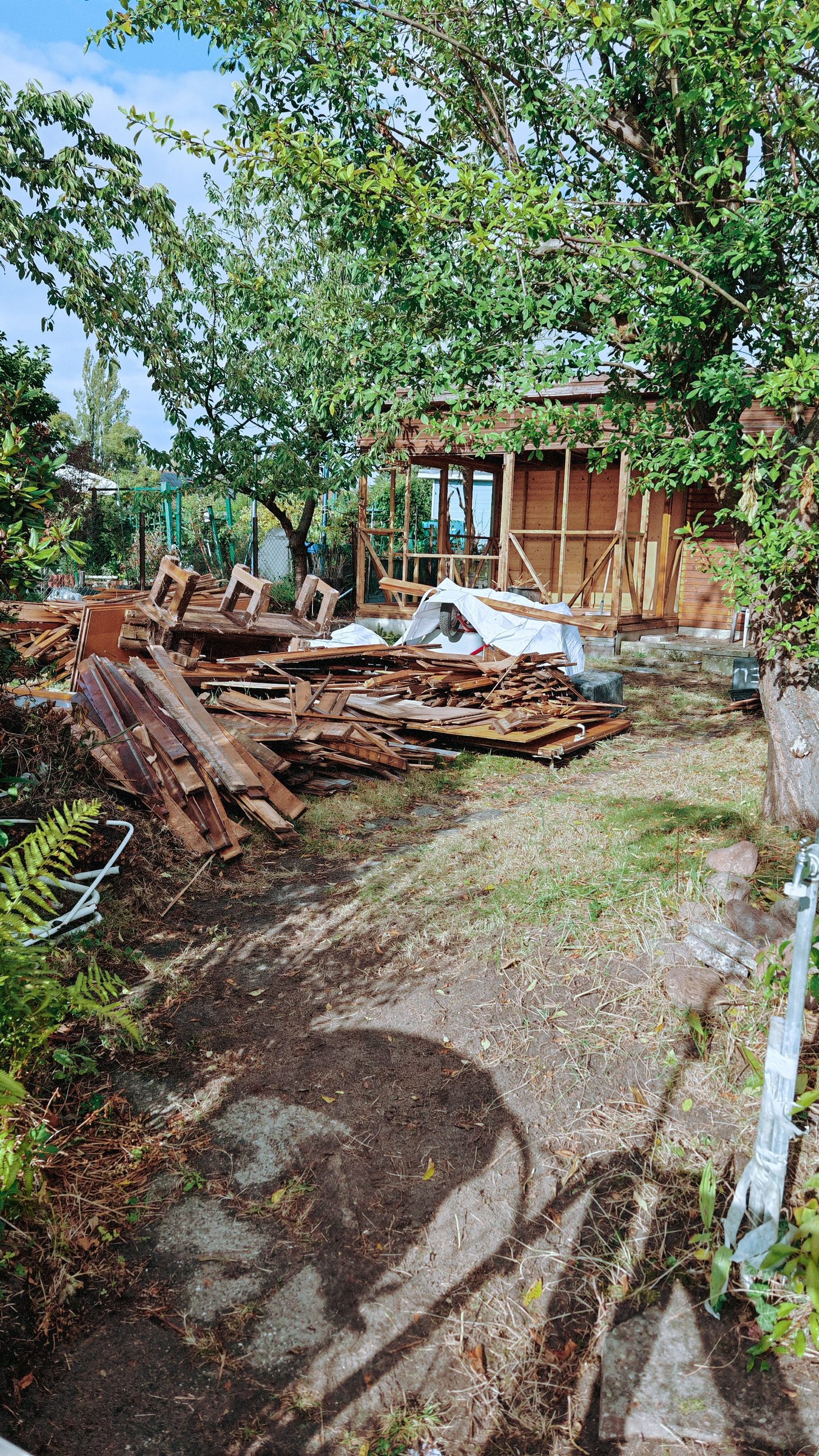 Gestapelte Bretter und Balken neben dem Gartenweg, im Hintergrund das Gartenhaus im Rückbau